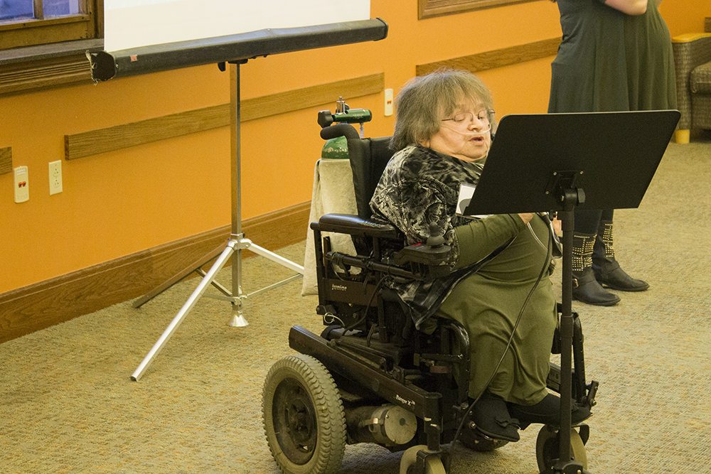 A woman in a wheelchair reads from a podium while talking into a microphone.